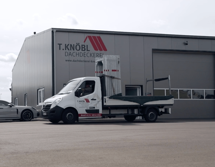 Weißer Transporter mit Schilder und T. KNÖBL Dachdeckerei Logo vor grauem Firmengebäude.