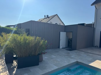 Braunes Gartenhaus mit offener Tür und weißem Rahmen, daneben ein Pool und eine große Pflanze in einem schwarzen Topf.