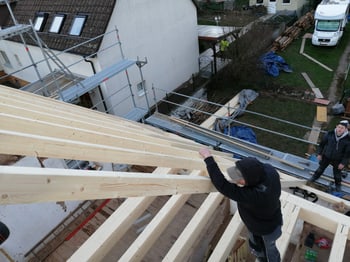 Zwei Bauarbeiter montieren Holzbalken auf einem Dach, ein Gerüst steht am Gebäude.