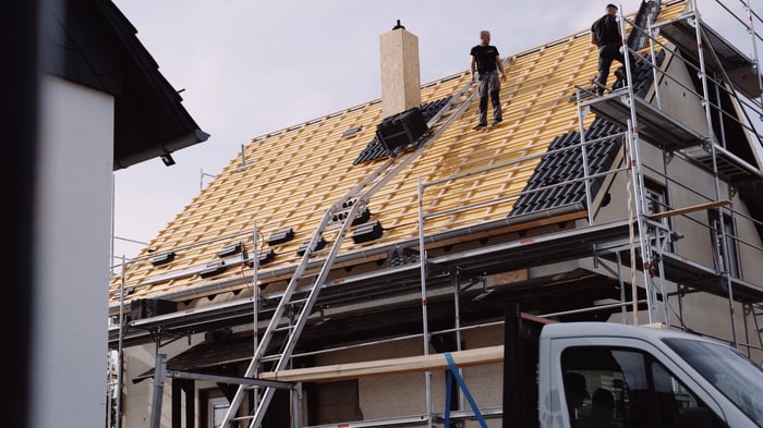Zwei Dachdecker auf einem Hausdach mit Holzlattung und teilweise verlegten Solarmodulen, Gerüst und Lieferwagen.
