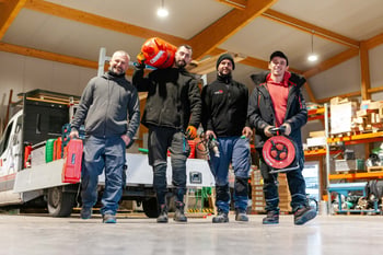 Vier Männer in Arbeitskleidung tragen Werkzeuge in einer Lagerhalle, mit einem weißen Transporter im Hintergrund.