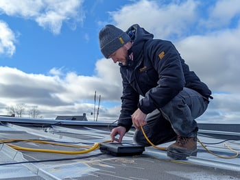 Ein Mann in Winterkleidung hockt auf einem Metallwellblechdach und bedient ein elektronisches Gerät mit gelben Kabeln.
