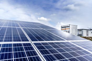 Solarmodule unter blauem Himmel mit einigen Wolken; im Hintergrund sind zwei weiße Container zu sehen.